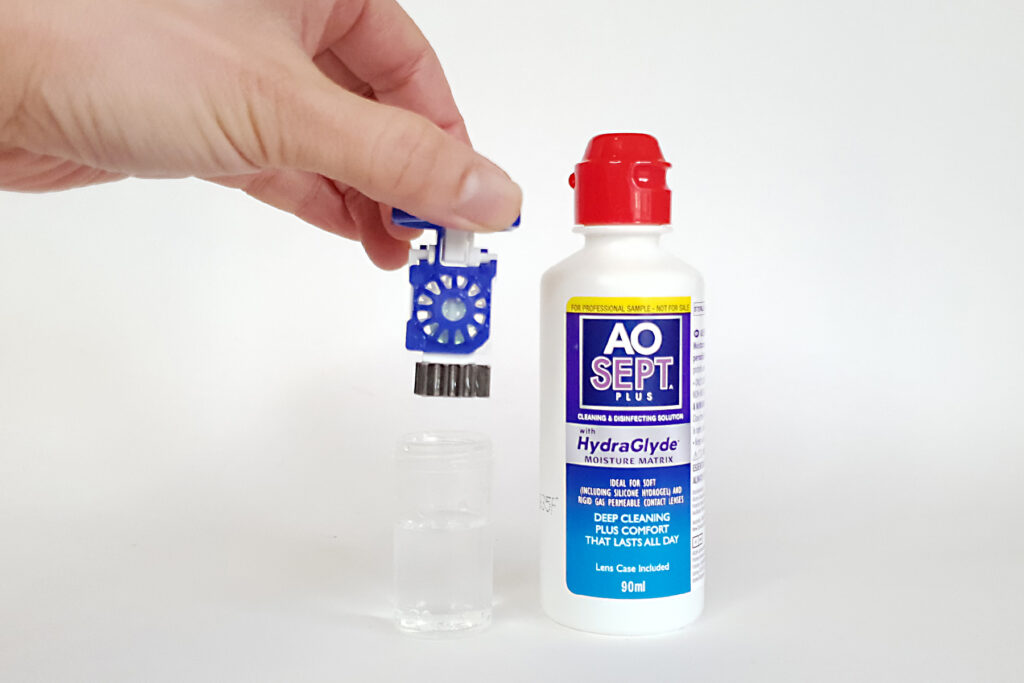 A hand inserting the OrthoK lenses inside its case with a bottle of AO Sept next to it